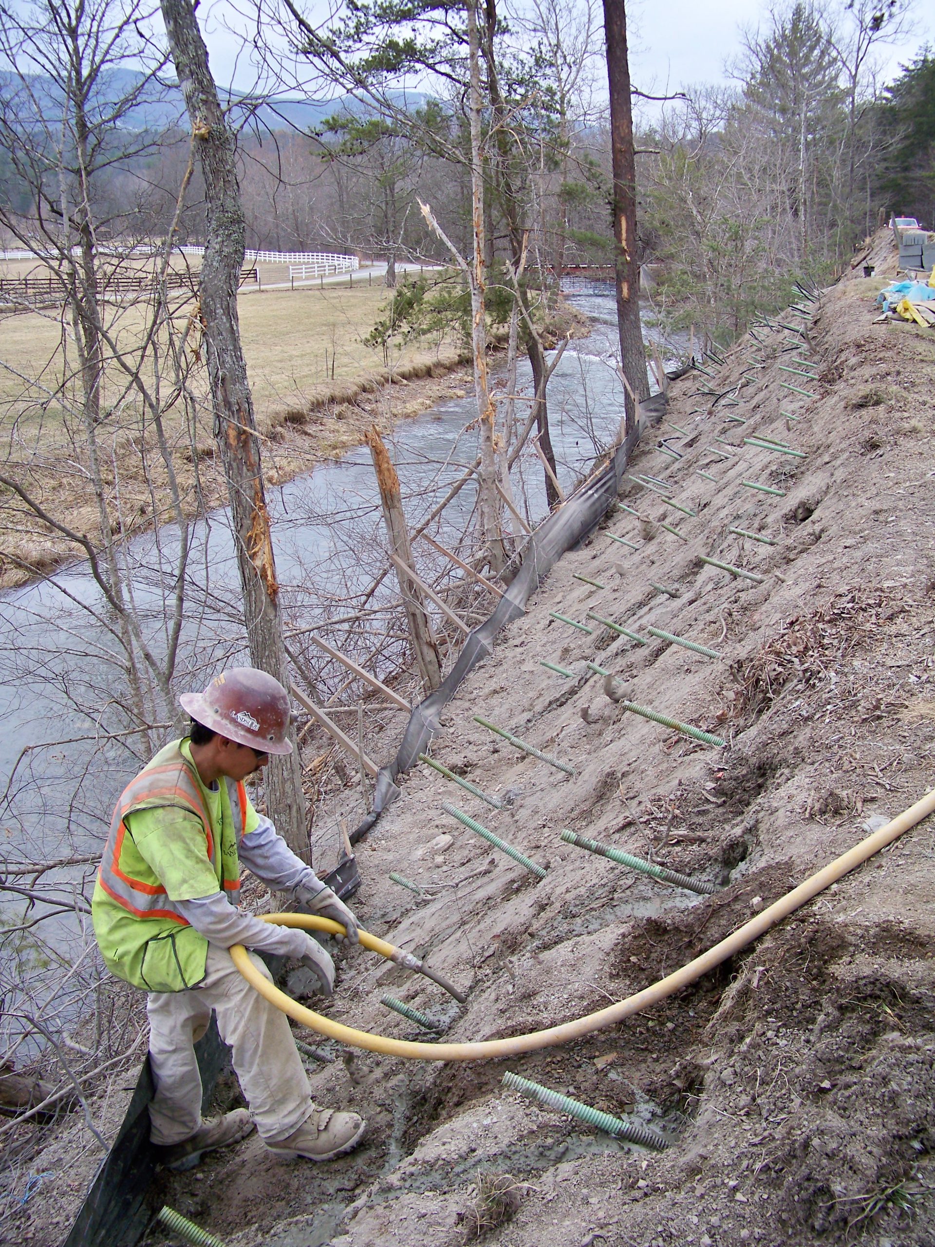 Worker installing soil nails and hoses on a steep slope above a riverbank for erosion control or slope stabilization.