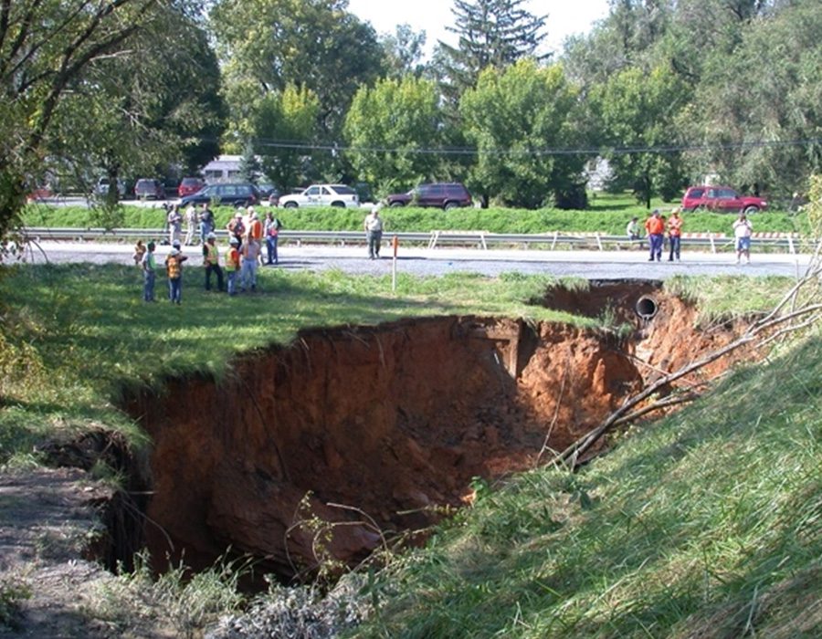 Large sinkhole near a road with people observing from a distance.