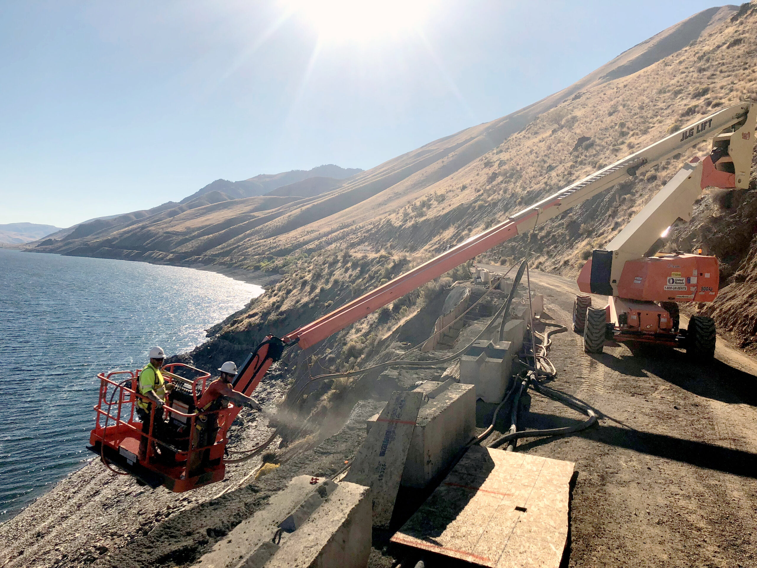 Two workers in a lift basket performing slope stabilization work above a river.