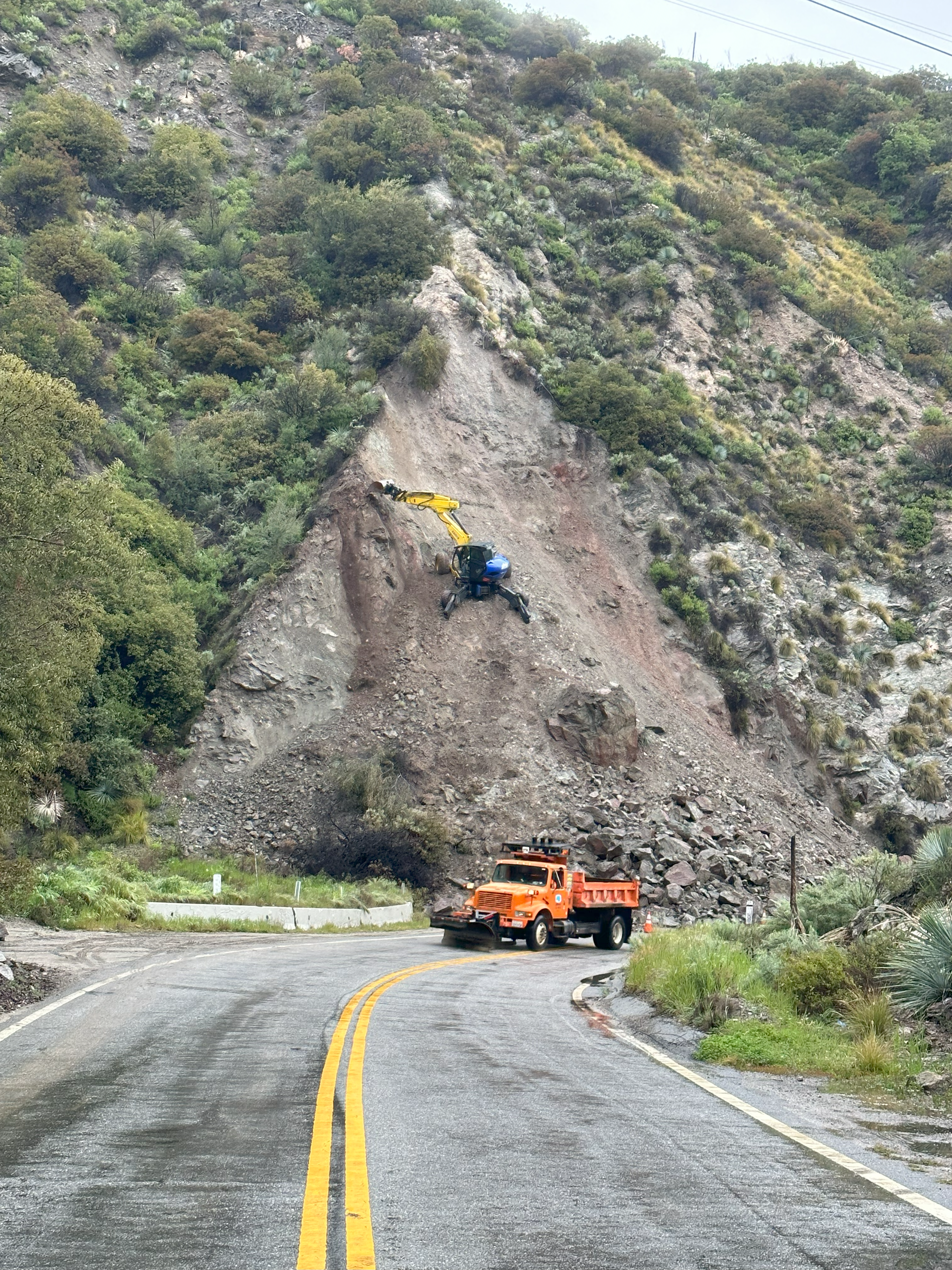 Spider excavator scaling a steep roadside slope with loose debris.