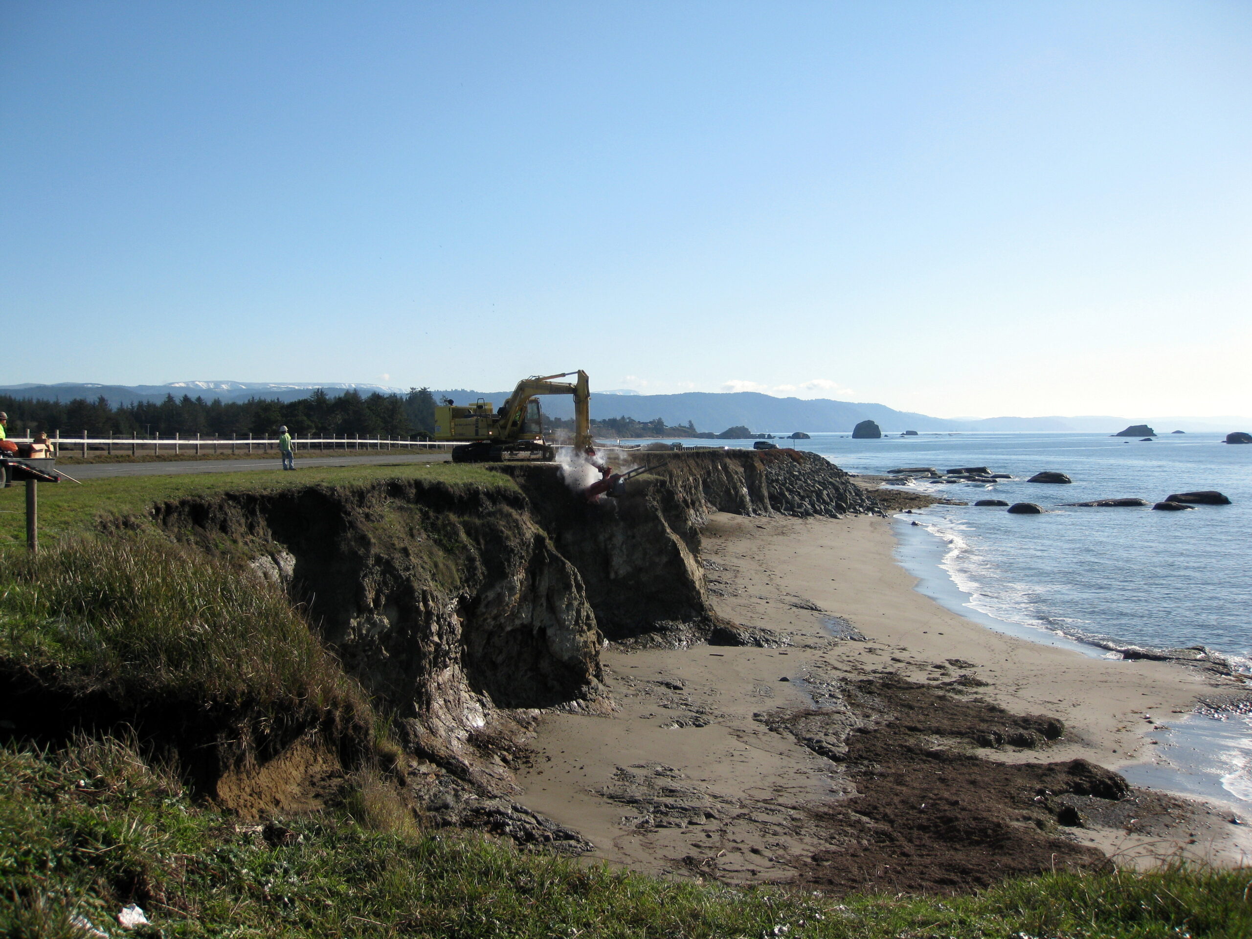 Excavator working near eroded coastal bluff above a sandy shoreline.