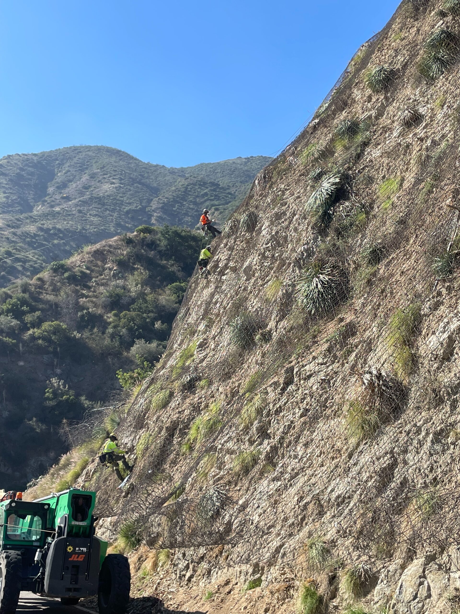 Crew members secured by ropes install draped rockfall mesh on a steep, rocky slope above a roadway.