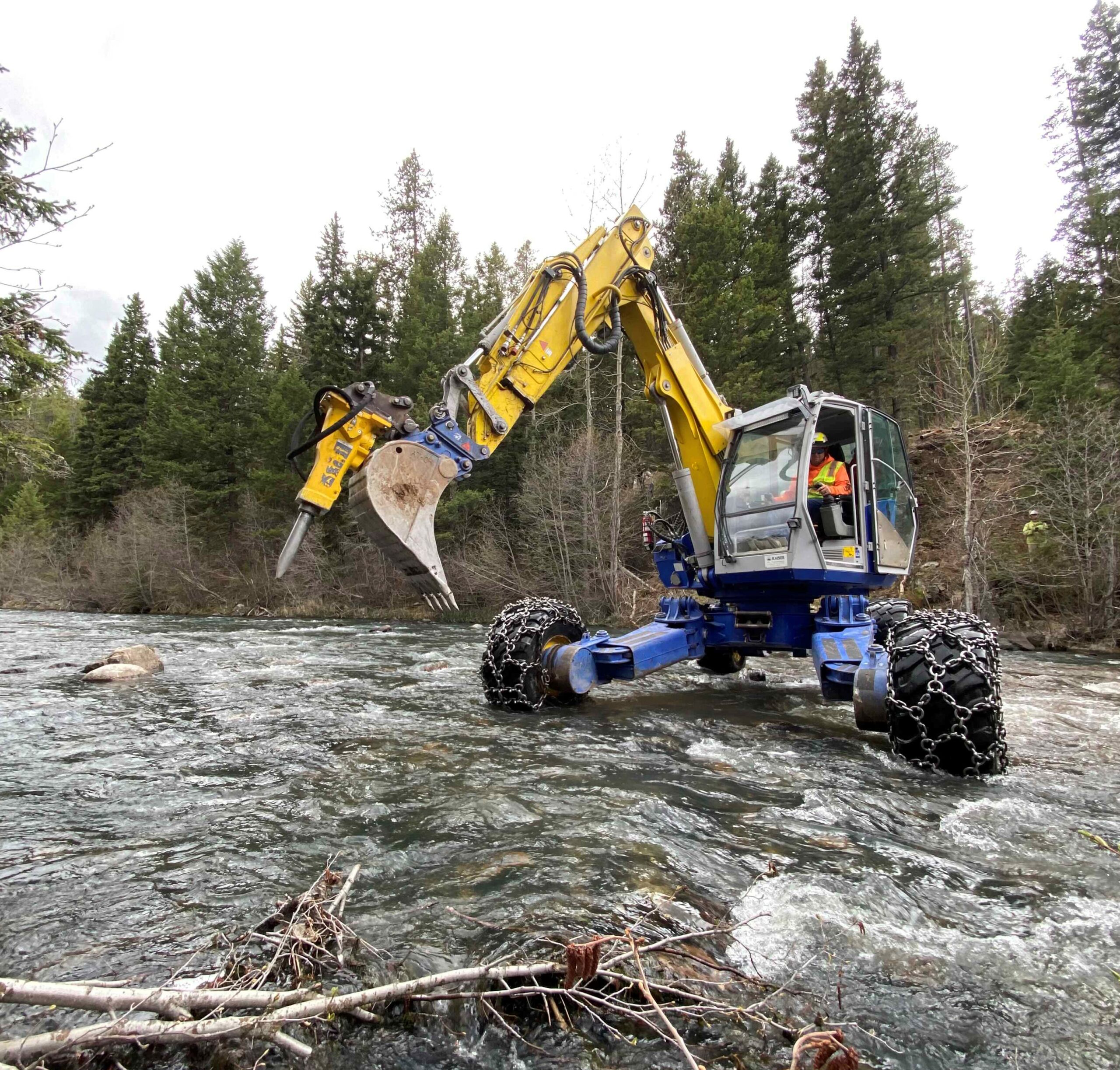 A spider excavator with chained wheels crossing a river, operated by a worker in a forested area.