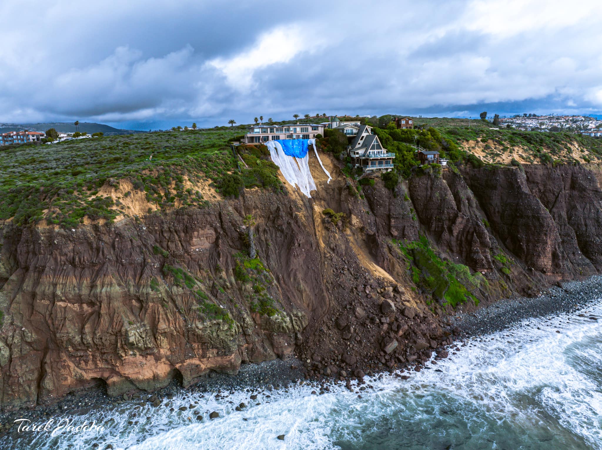 Oceanfront bluff with landslide damage covered by tarps below cliffside homes.