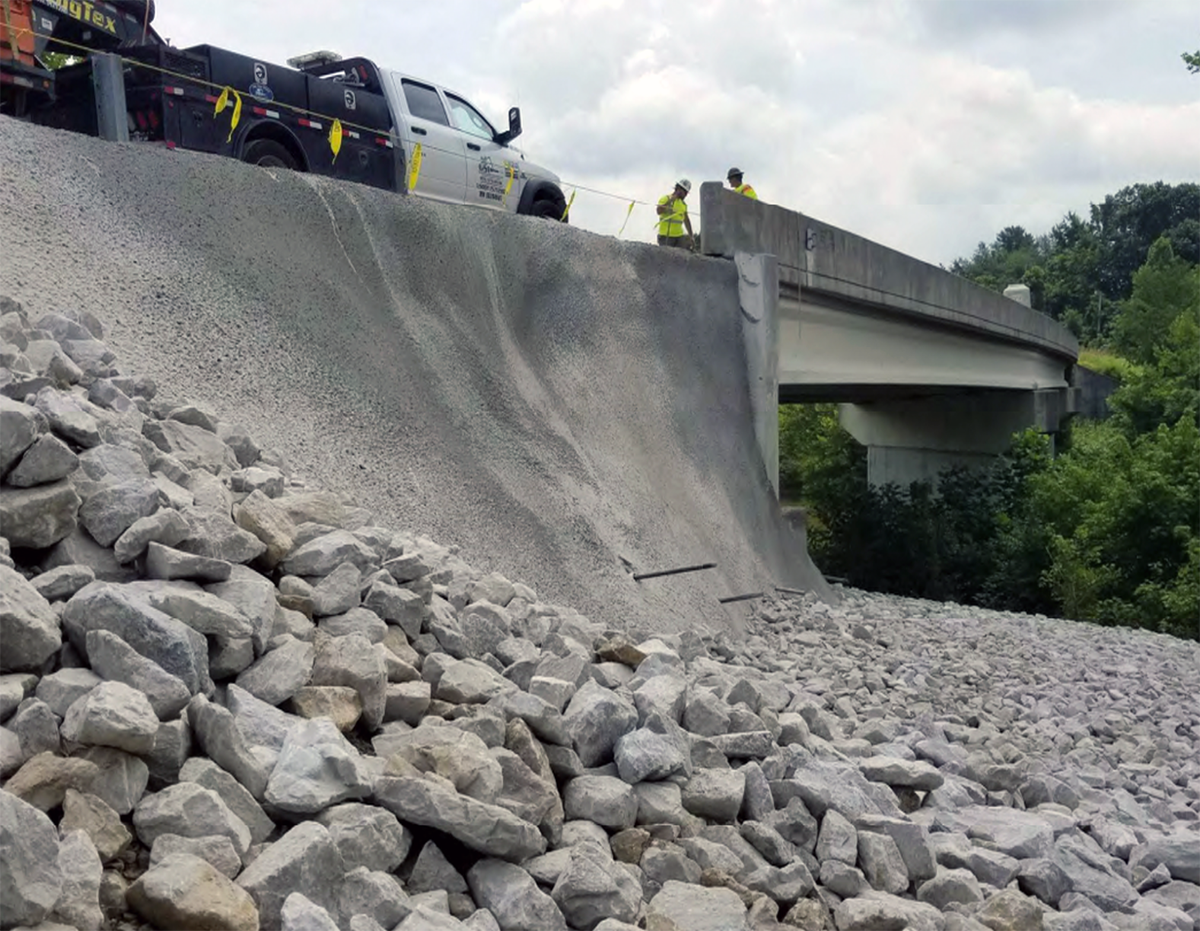 Freshly placed rock riprap at the base of a concrete embankment wall beneath a bridge.