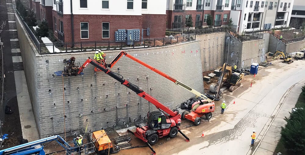 Crews using lift equipment to install soil nails on a tall retaining wall near residential buildings.