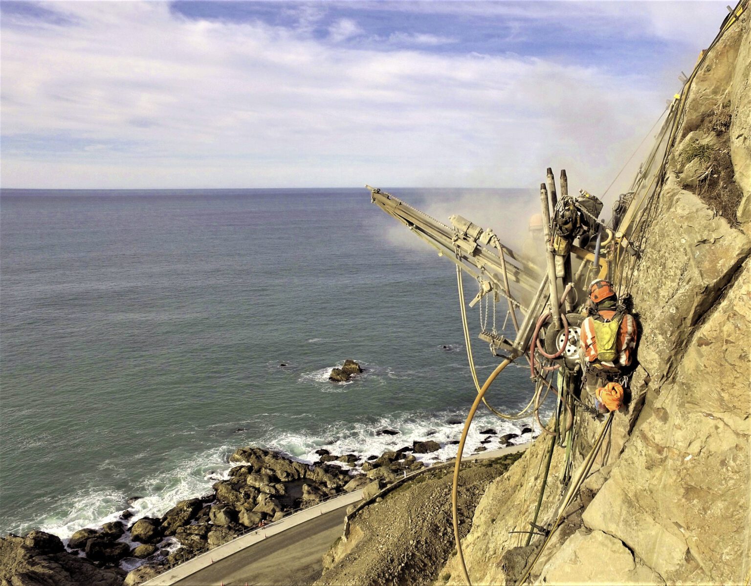 Worker operating drilling equipment on a steep coastal cliff.