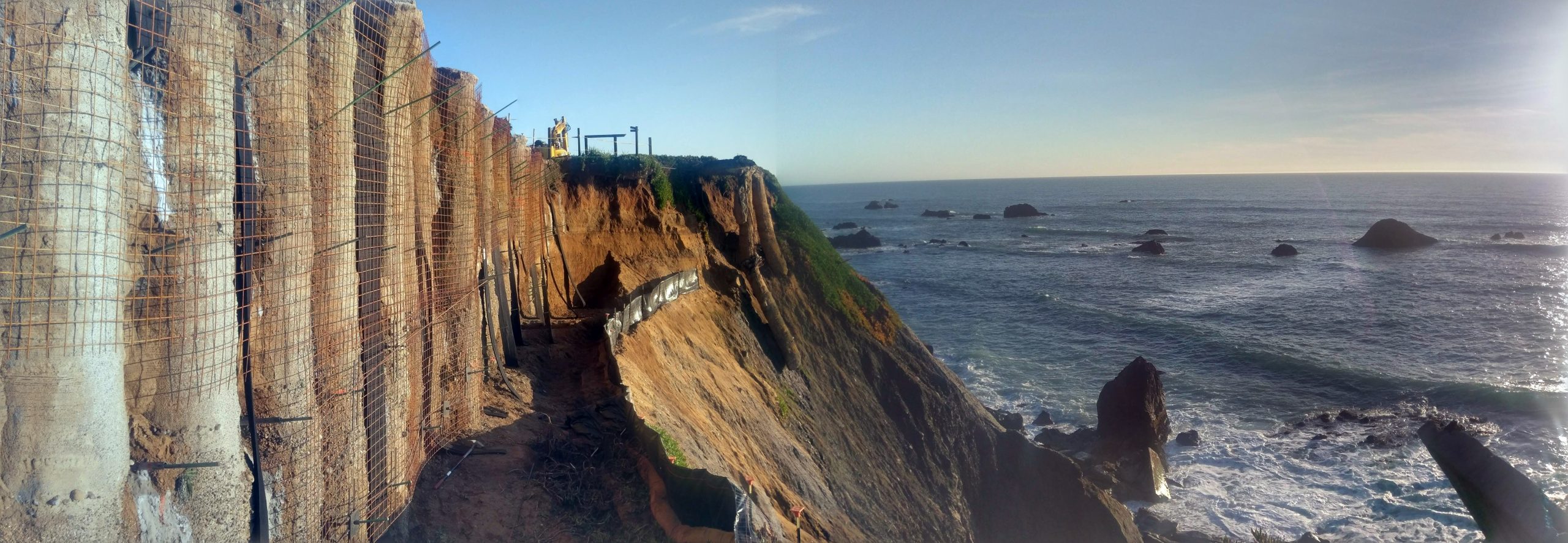 Coastal cliff stabilization project with soil nails and wire mesh installed along a vertical cliff face overlooking the ocean.