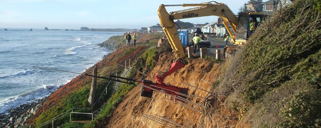 Excavator and crew performing stabilization work on an eroding coastal bluff above the ocean.