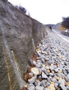 Shotcrete wall with drainage holes above a rock-covered slope next to a road.