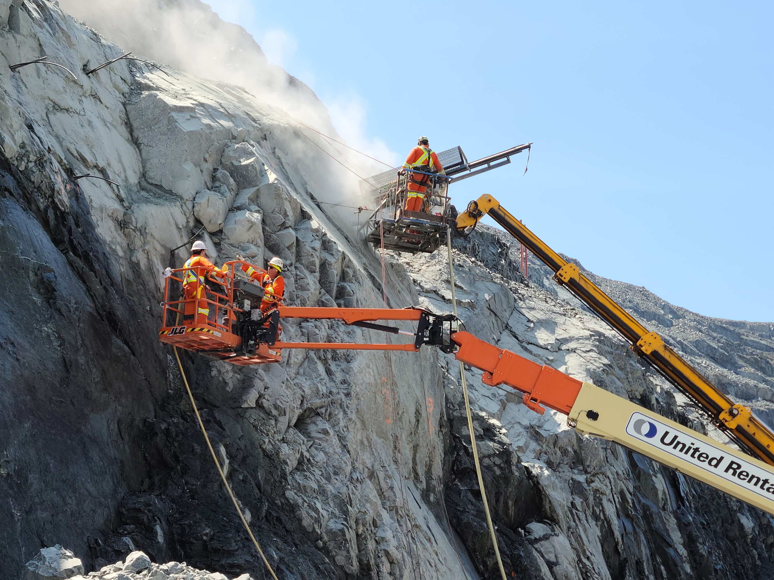 Workers in lift platforms drilling into a steep rock face for rockfall mitigation.