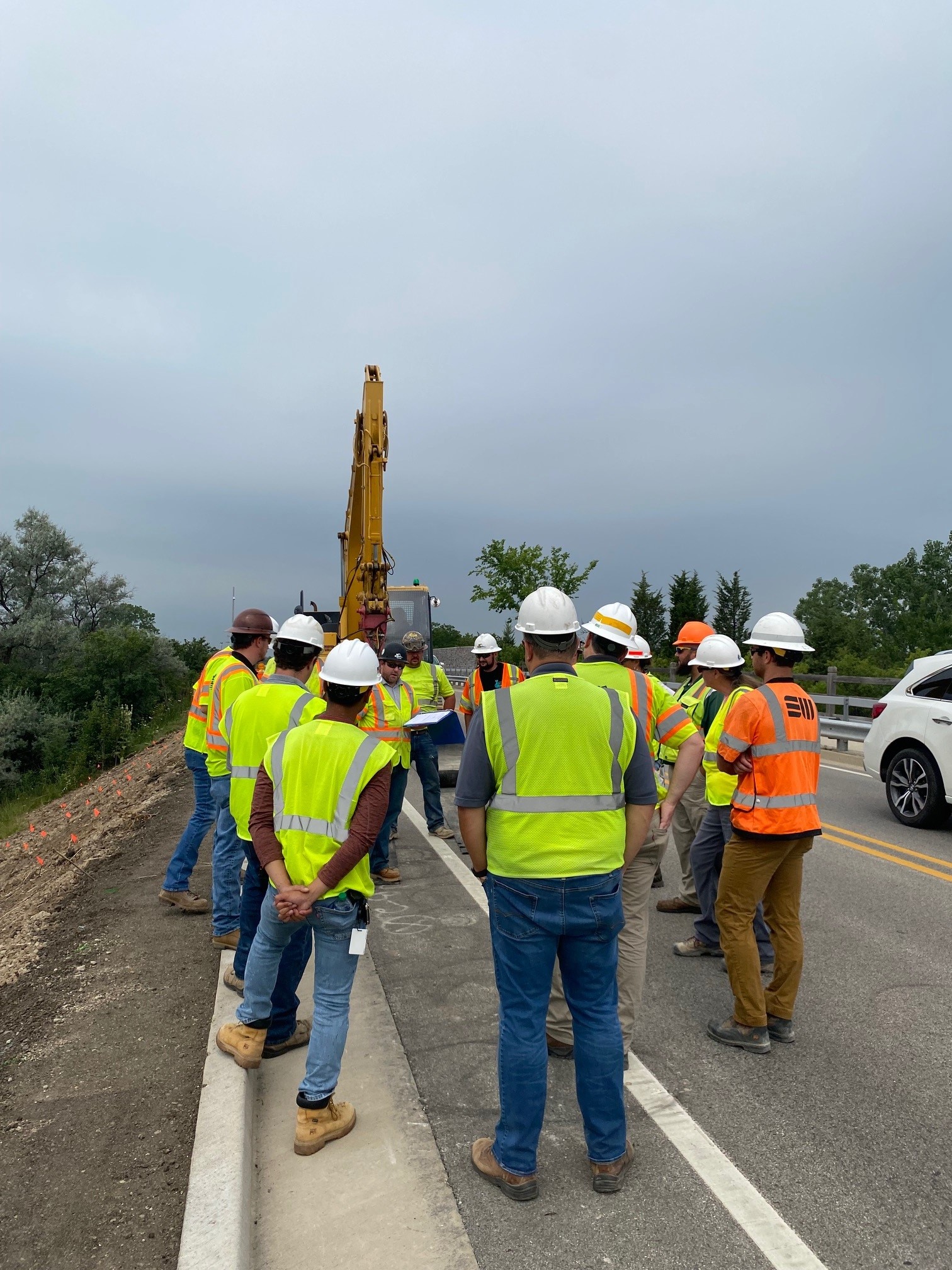 Construction crew in safety vests and hard hats gathered for a roadside safety briefing.