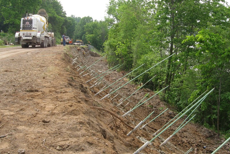 Center rebar installation along a roadside slope, with angled soil nails in place and construction vehicles in the background.