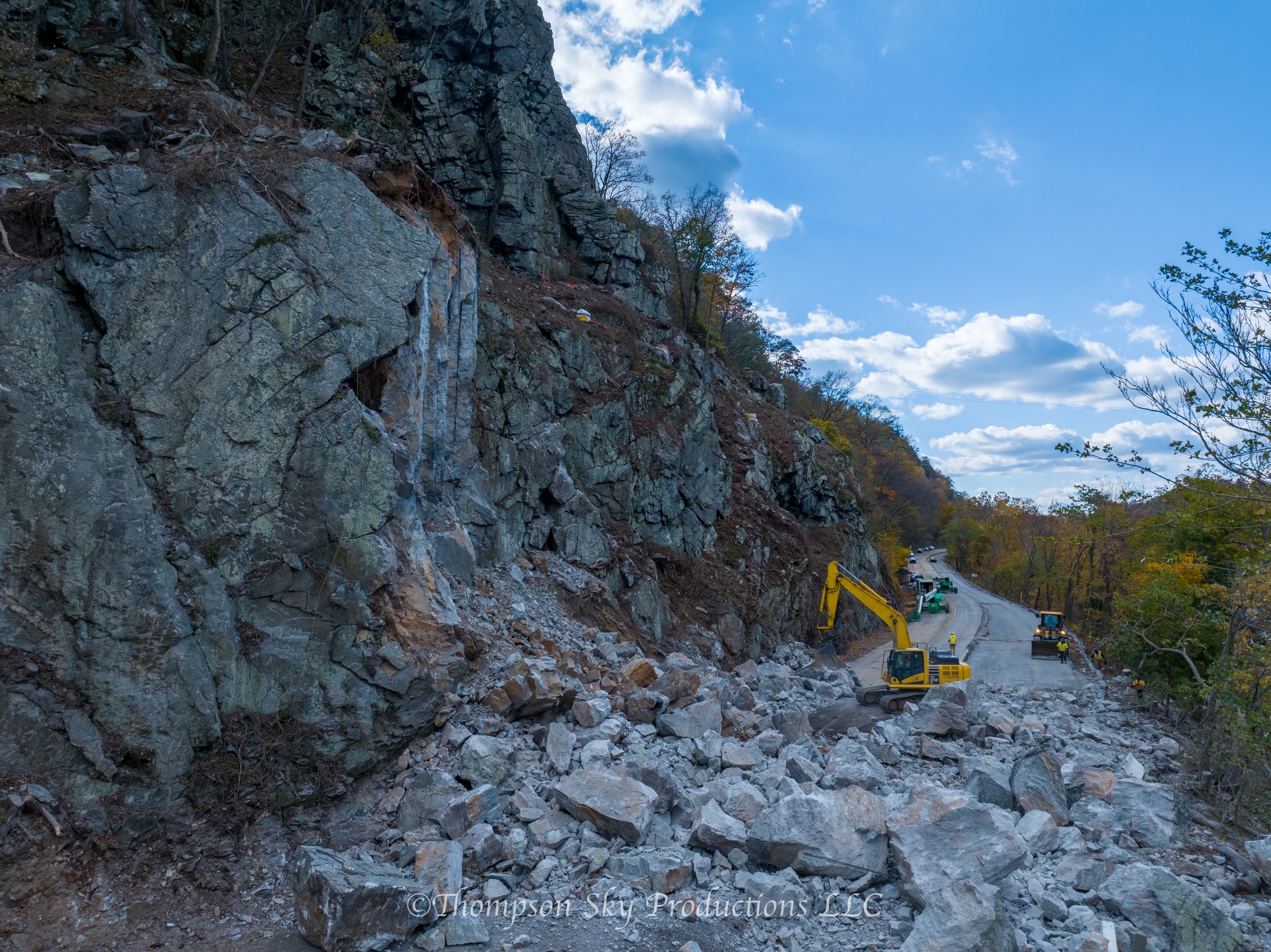 Excavator clears large rockfall debris from a roadway beneath a steep cliff.