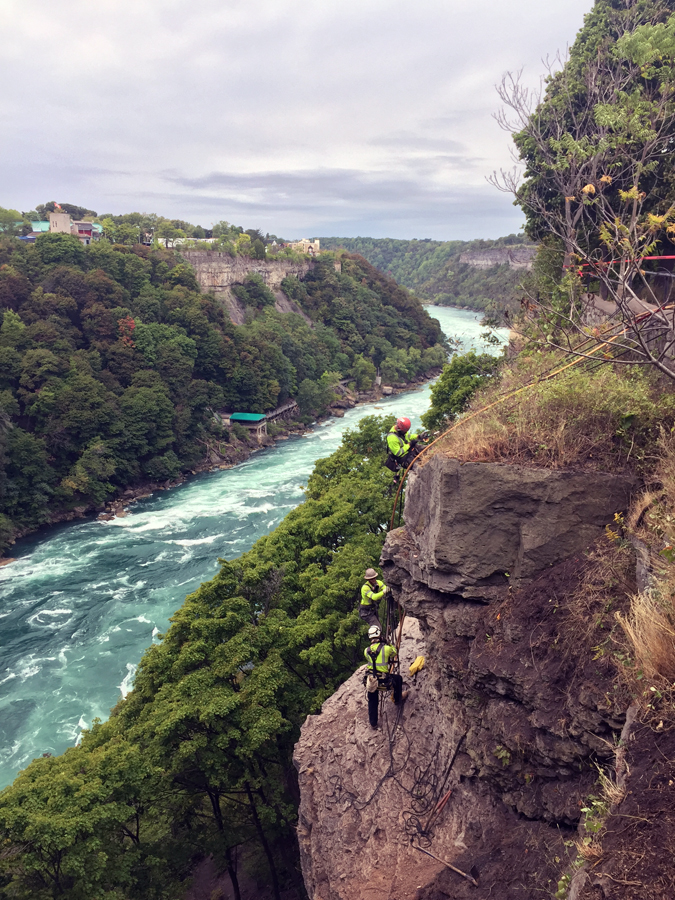 Workers perform rope access rock scaling above a river gorge.