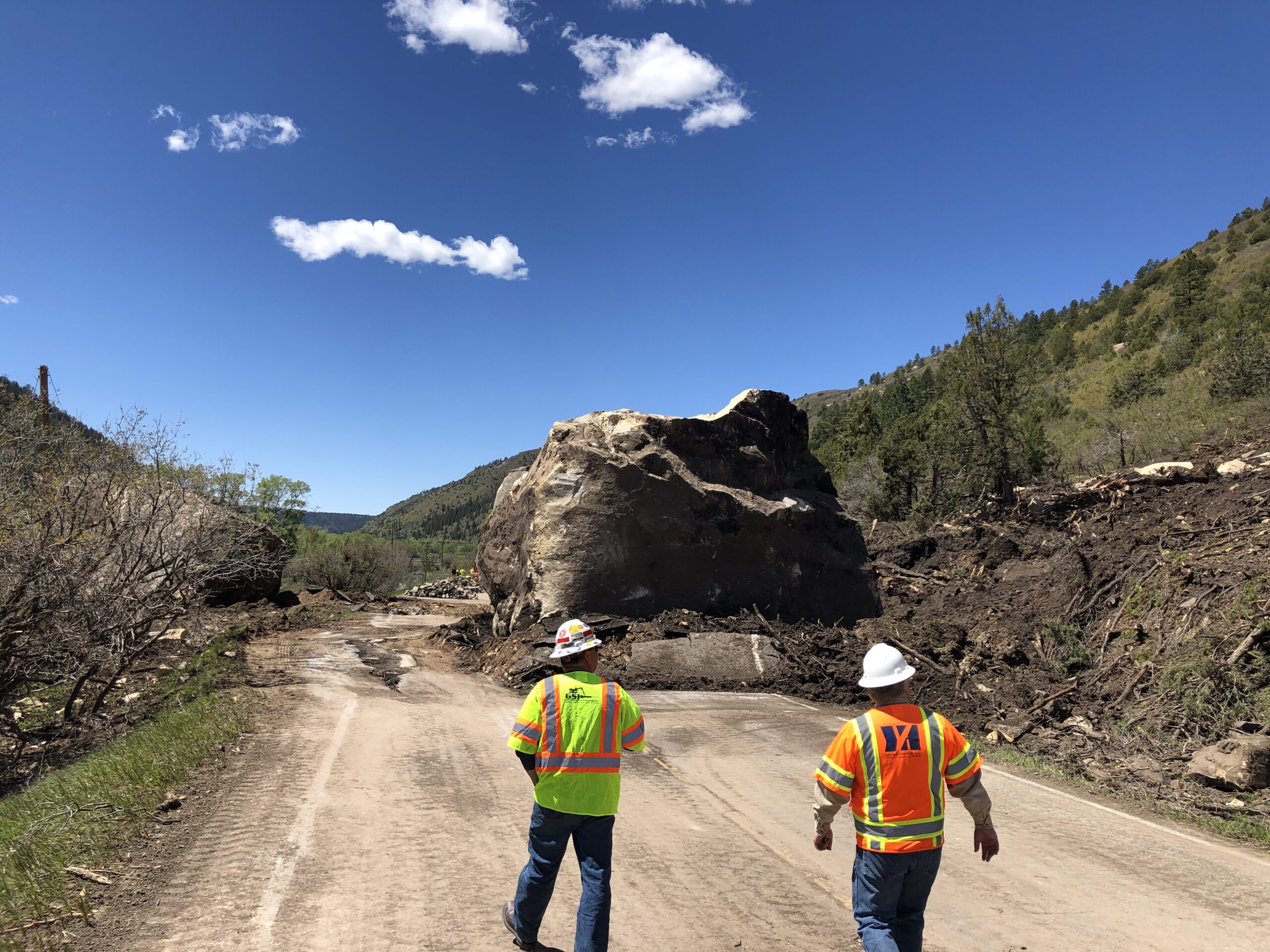 Two workers approach a massive boulder blocking a dirt road.
