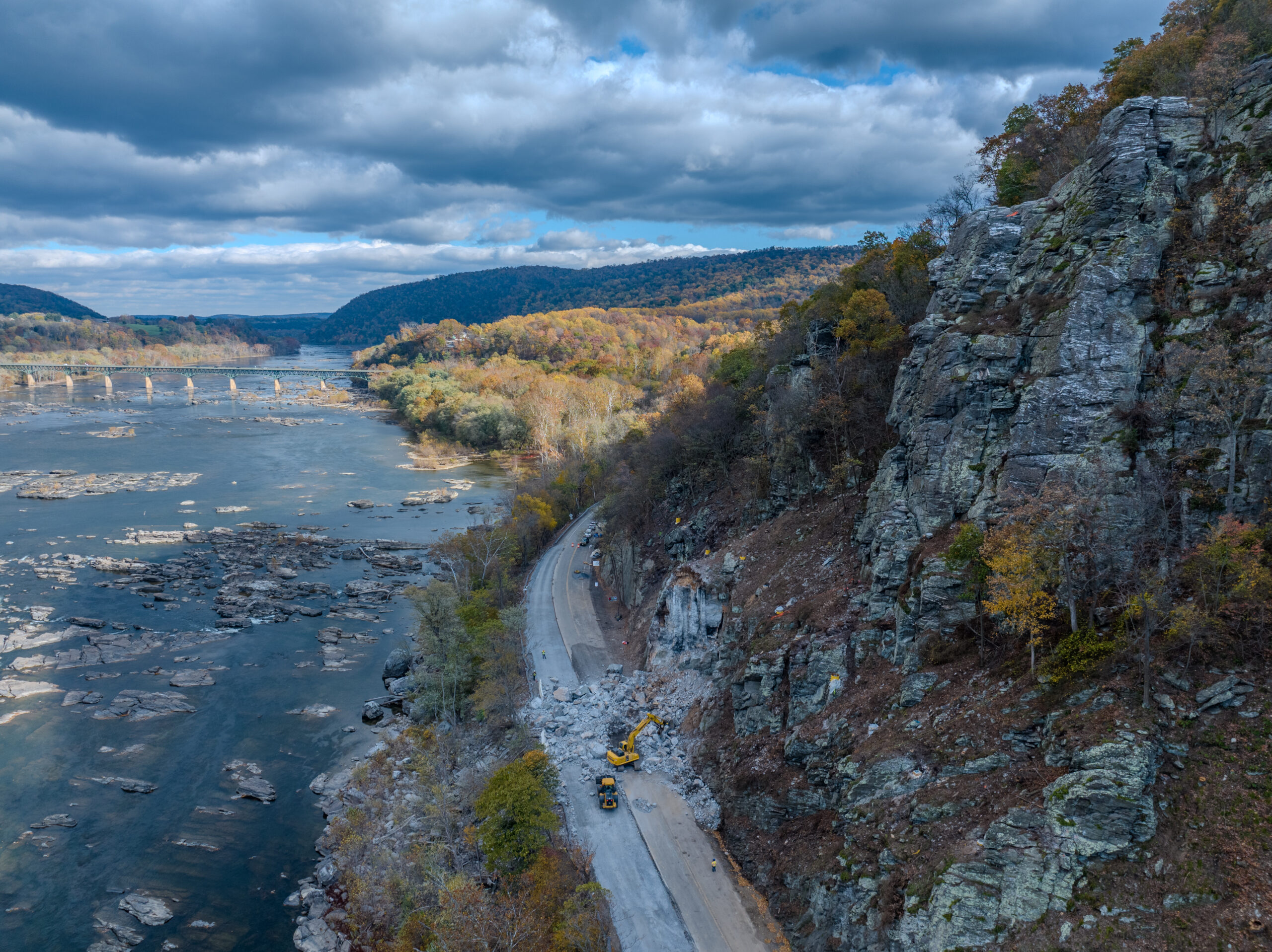 Excavator clears rock debris along a riverside road.
