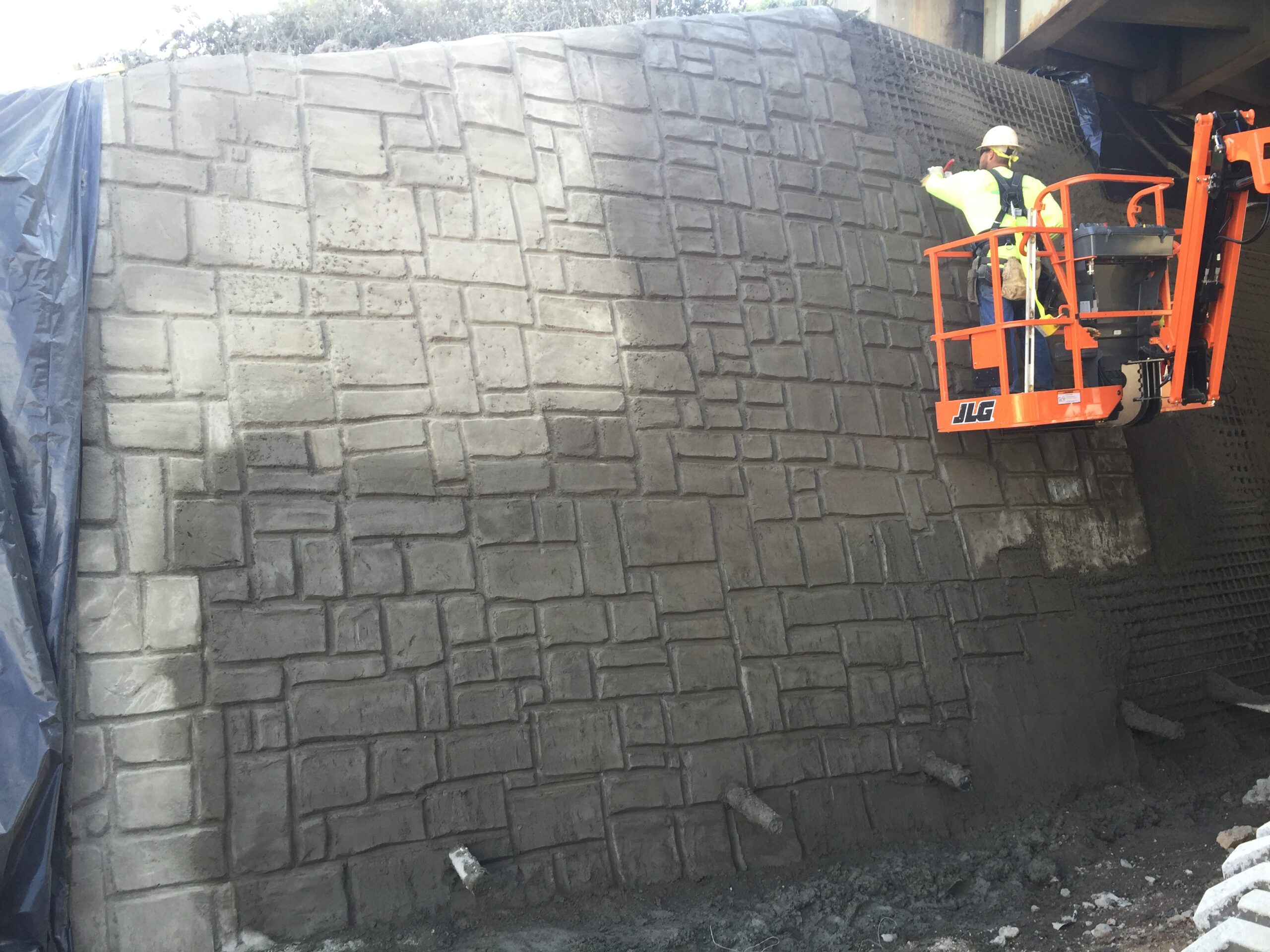 Worker on a lift applying stain to a stamped ashlar-pattern shotcrete wall.