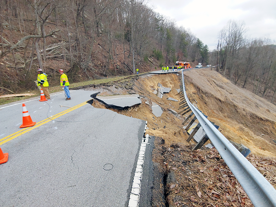 Roadway collapse due to embankment failure, with workers assessing damage and erosion near a steep slope.