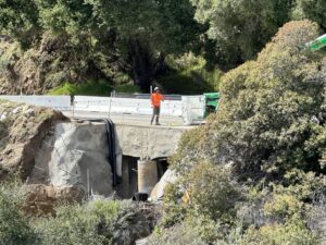 Worker in orange safety gear stands on a repaired roadside.