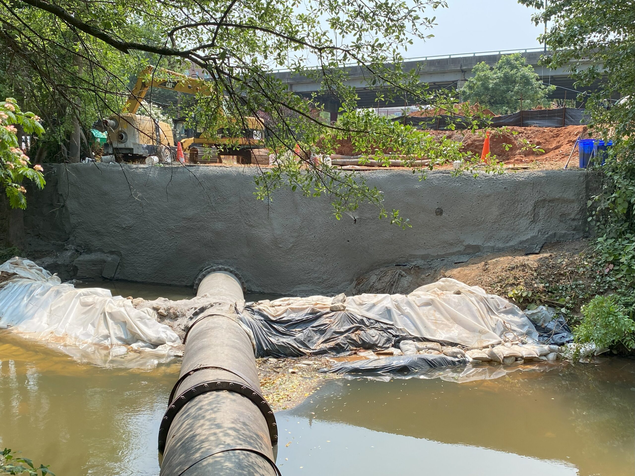 Construction site with shotcrete wall and large pipe crossing a stream.