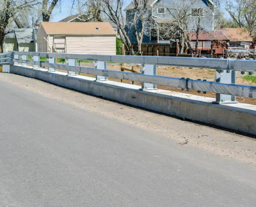 Galvanized w-beam bridge rail on concrete curb alongside rural road with residential backdrop