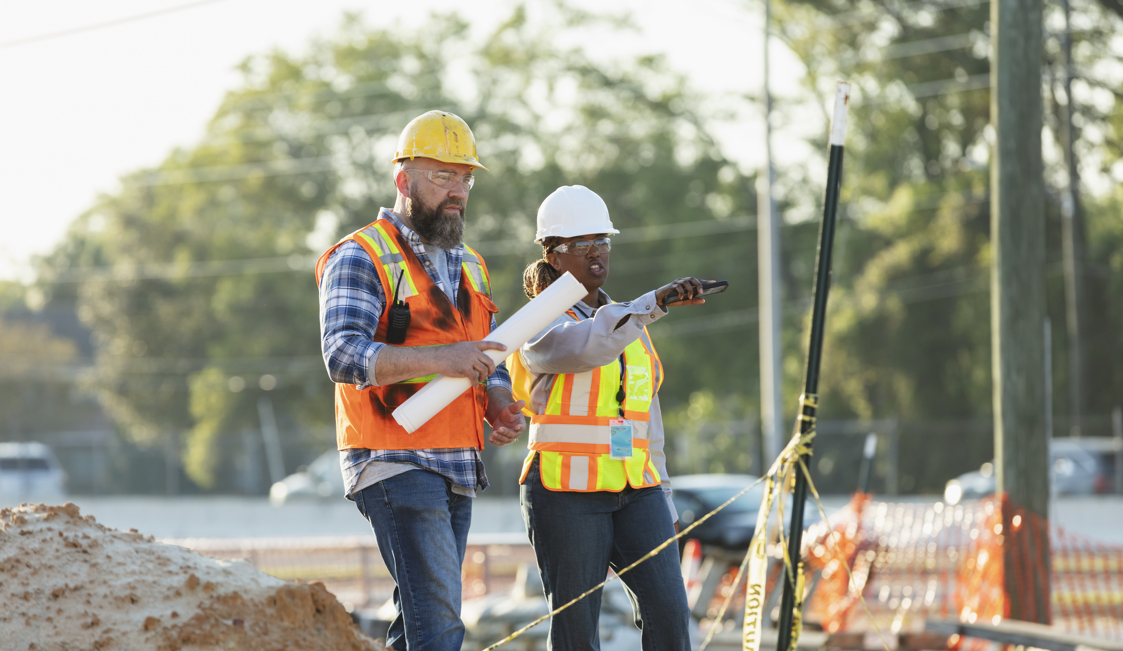 Two highway construction project managers in hard hats and safety vests reviewing plans at a job site