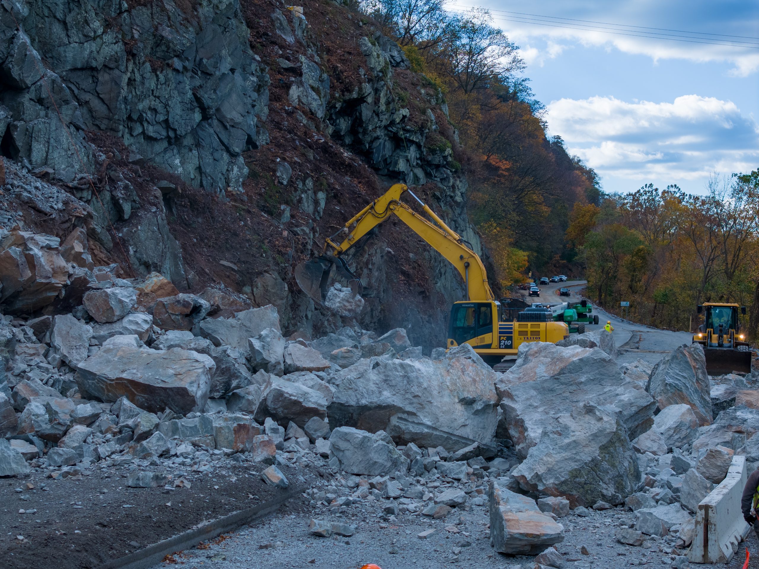 Excavator clearing large boulders from a rockfall along a mountain road.