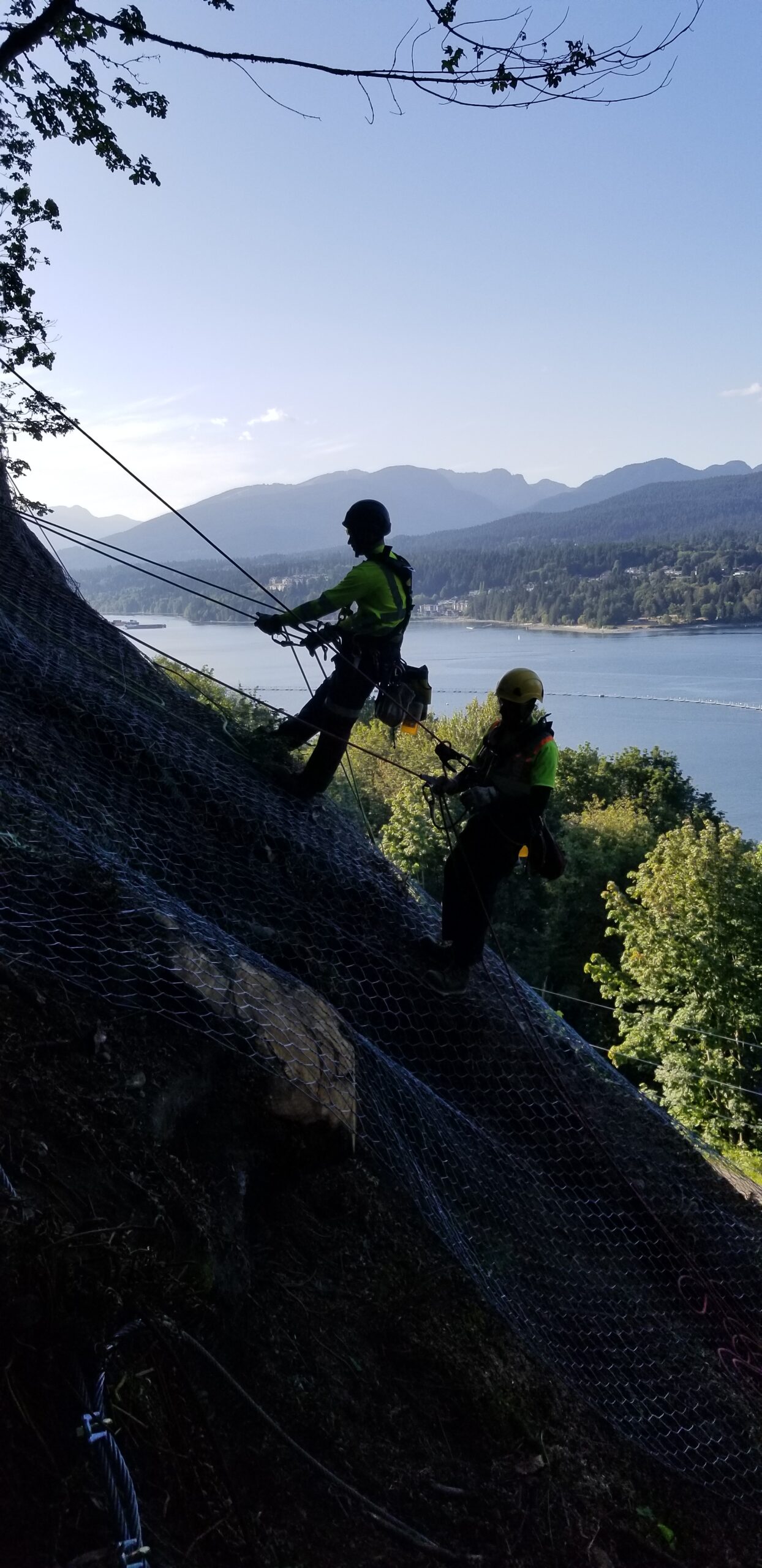 Workers in safety gear rappel down a steep, mesh-covered rock slope during a rockfall mitigation project, with a lake and mountains in the background.