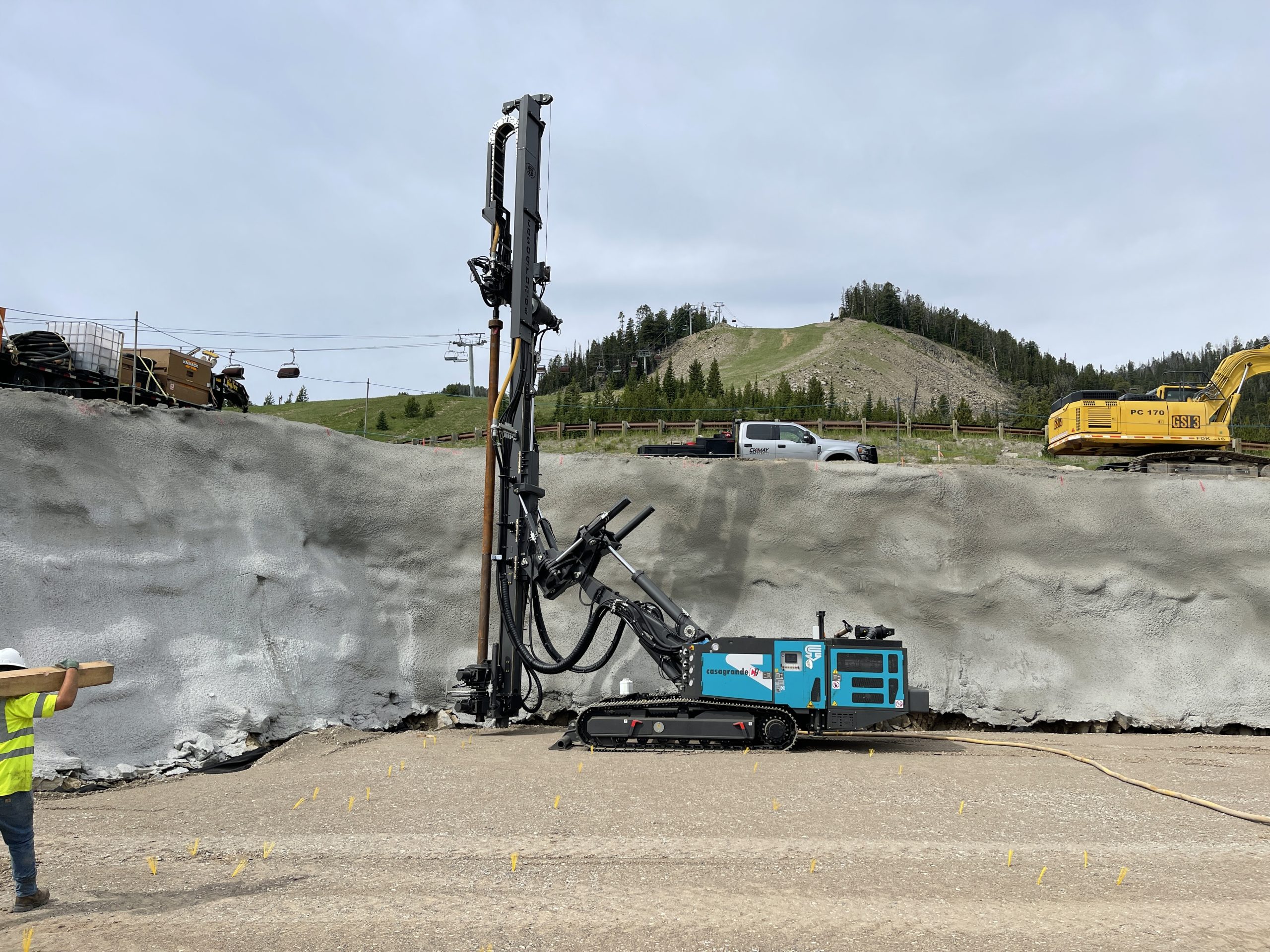 Drill rig installing soil nails into a shotcrete-covered slope beside a road.