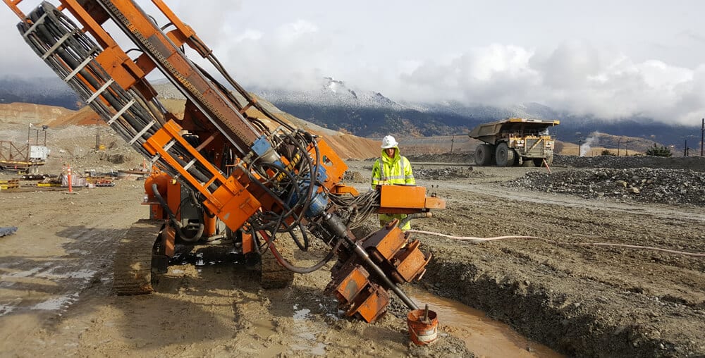 Worker operating orange drilling equipment for grouting on a muddy construction site.