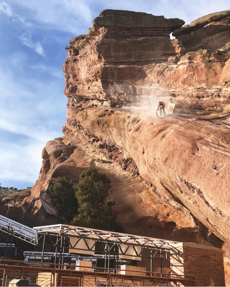 Steep red rock cliff with protective mesh installation above a concert stage structure.