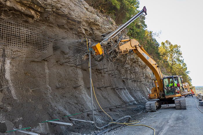 Excavator applying shotcrete to stabilize a roadside slope.