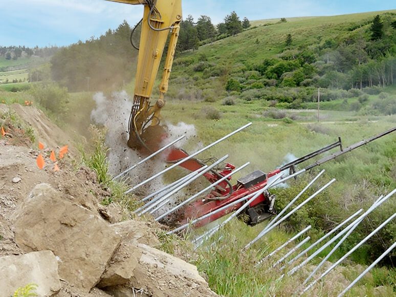 Excavator-mounted drill installing soil nails into a steep grassy slope.