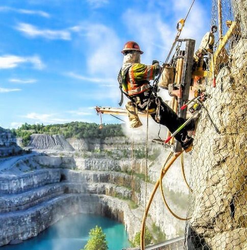 Worker suspended by ropes drilling into a rock face above a quarry.