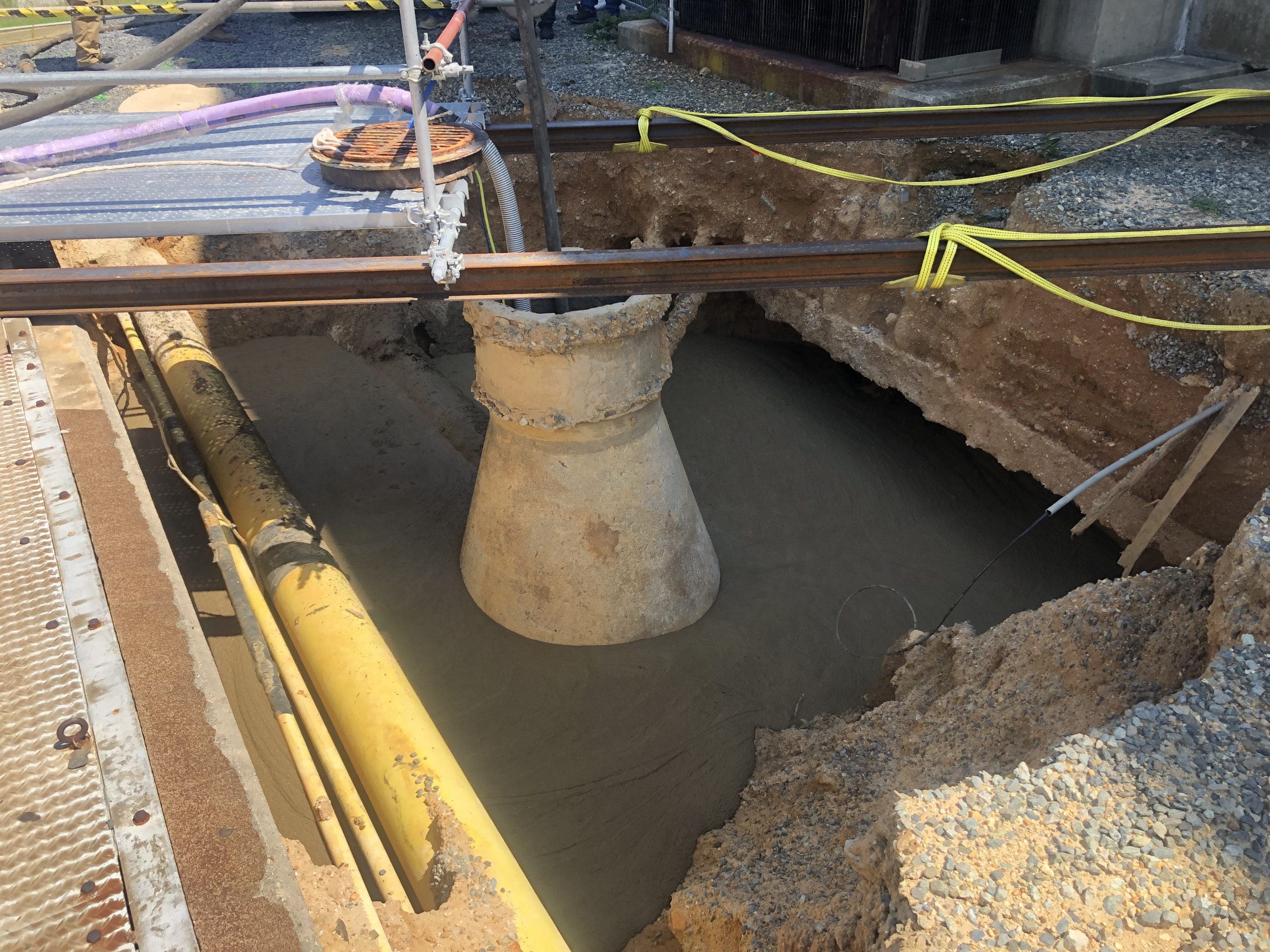 Construction site showing sinkhole repair with concrete grouting around a large pipe and exposed utility lines.