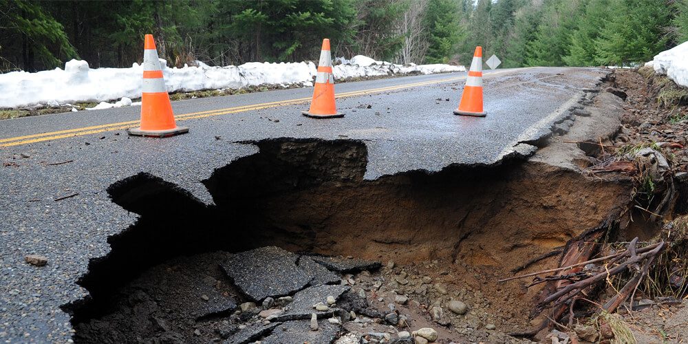 Partially collapsed roadway with a large sinkhole and traffic cones placed for safety in a snowy, forested area.